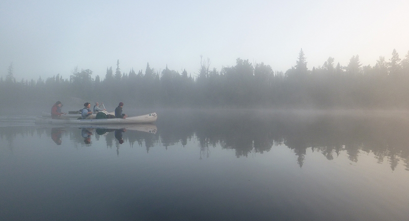 A canoe is paddled by outward bound students on very calm water. The air is foggy, and the water reflects the tree-lined shore. 